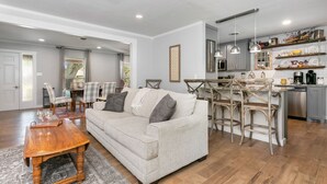 A modern living room with a sofa, wooden coffee table, and dining chairs. A kitchen with bar stools and pendant lights is visible in the background.
