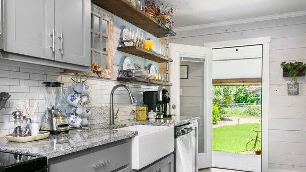 A modern kitchen with grey cabinets, a granite countertop, and a view of a garden through an open door.