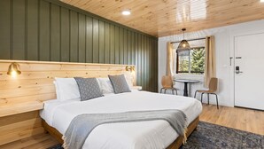 A bedroom with a wooden headboard, a large bed with white bedding, a patterned throw blanket, and a blue rug. There is a window with a view of greenery outside. - Standard Room w/ King Bed, Balcony, Fireplace, Mountain Views Room #13 (Lake Placid)