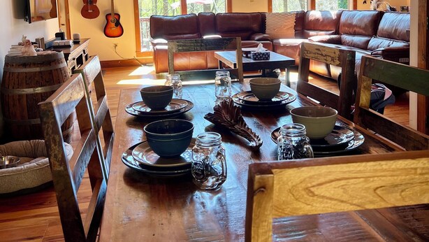 Un intérieur chaleureux et rustique, avec une table à manger en bois dressée pour le repas, un canapé en cuir et une grande fenêtre donnant sur la forêt.