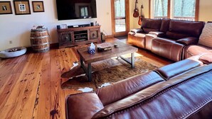 Un vaste salon doté d’un plafond en bois, de canapés en cuir, d’un téléviseur à écran plat et de grandes fenêtres offrant une vue sur les arbres.