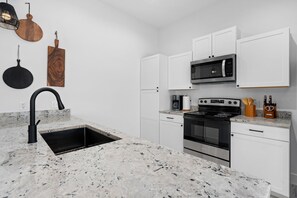 Kitchen features a sleek stove, stainless steel oven, and microwave above white cabinets, along with a mounted cutting board and sink.