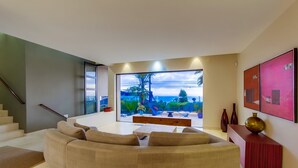 A modern living room with a curved sofa, a wooden coffee table, and a large window offering a view of greenery and a distant mountain.