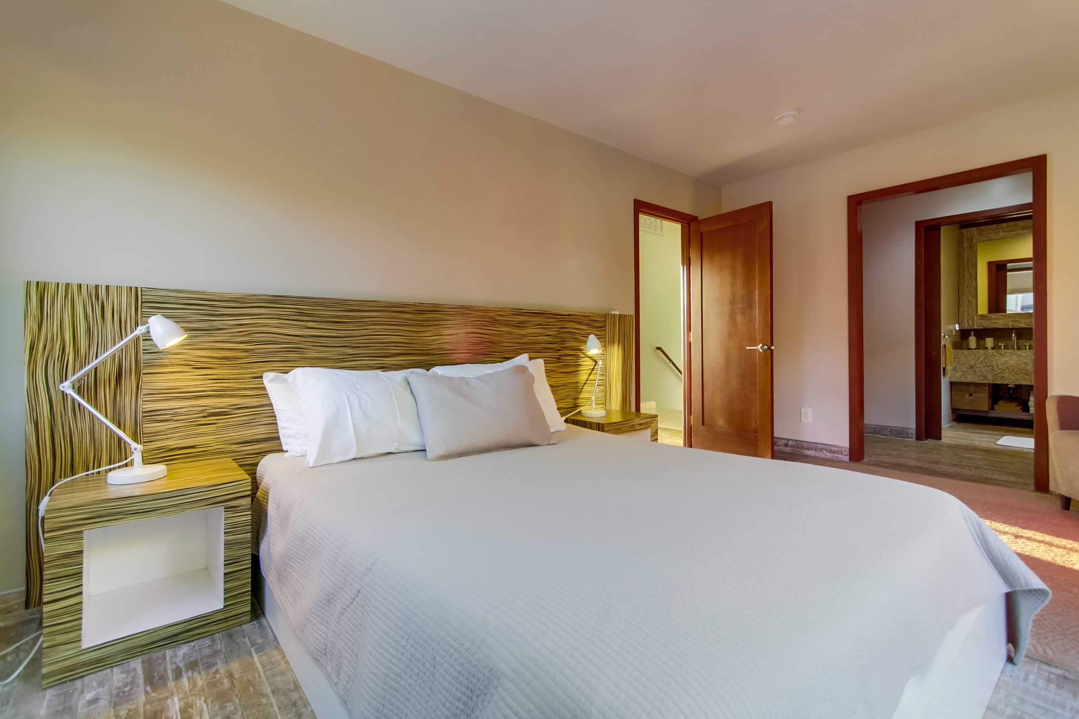 Habitación de hotel con una cama grande, dos mesitas de noche con lámparas, un cabecero de madera y un espejo sobre la cama.