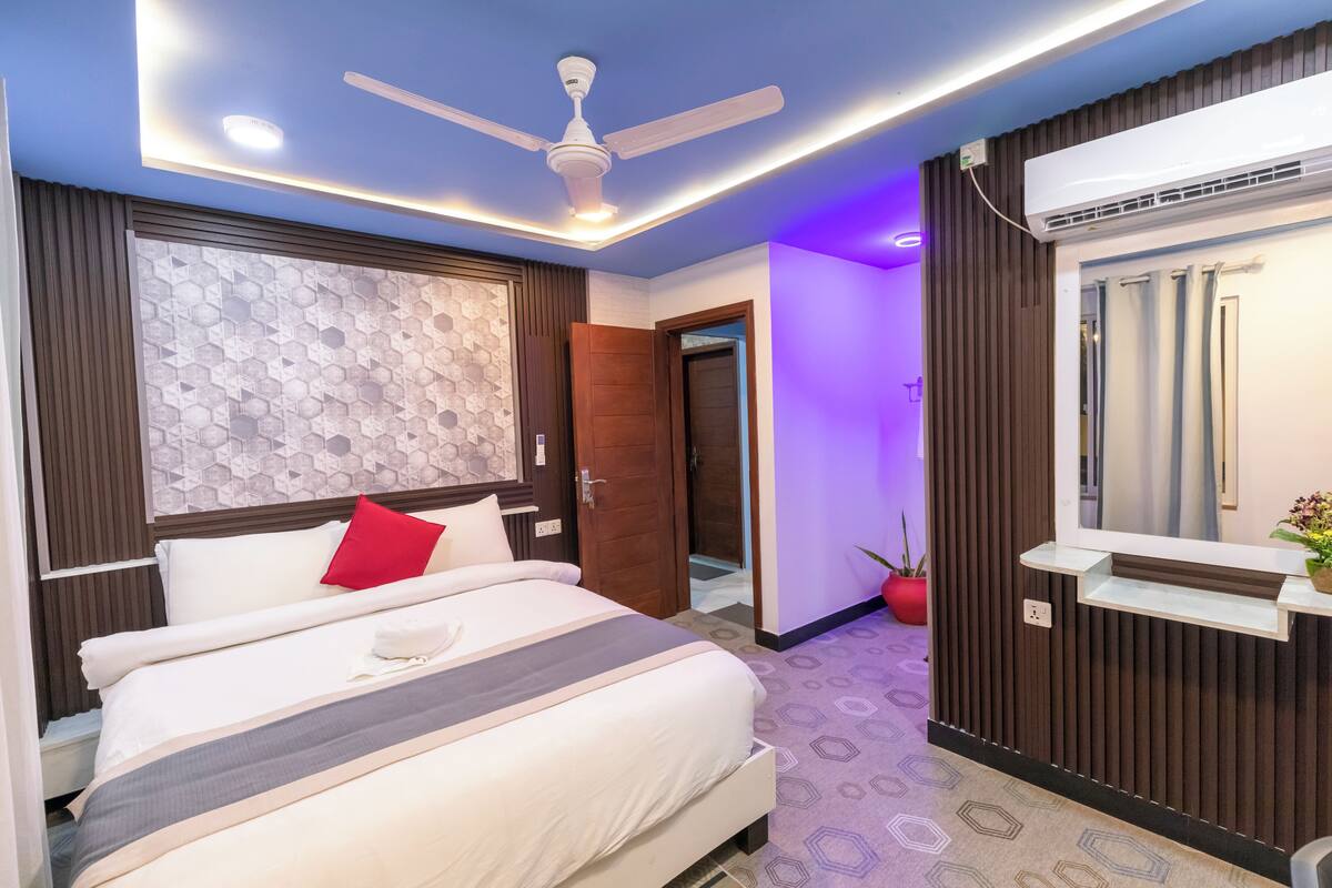 A hotel room with a bed, a ceiling fan, a wall-mounted air conditioner, and a small shelf with a potted plant.