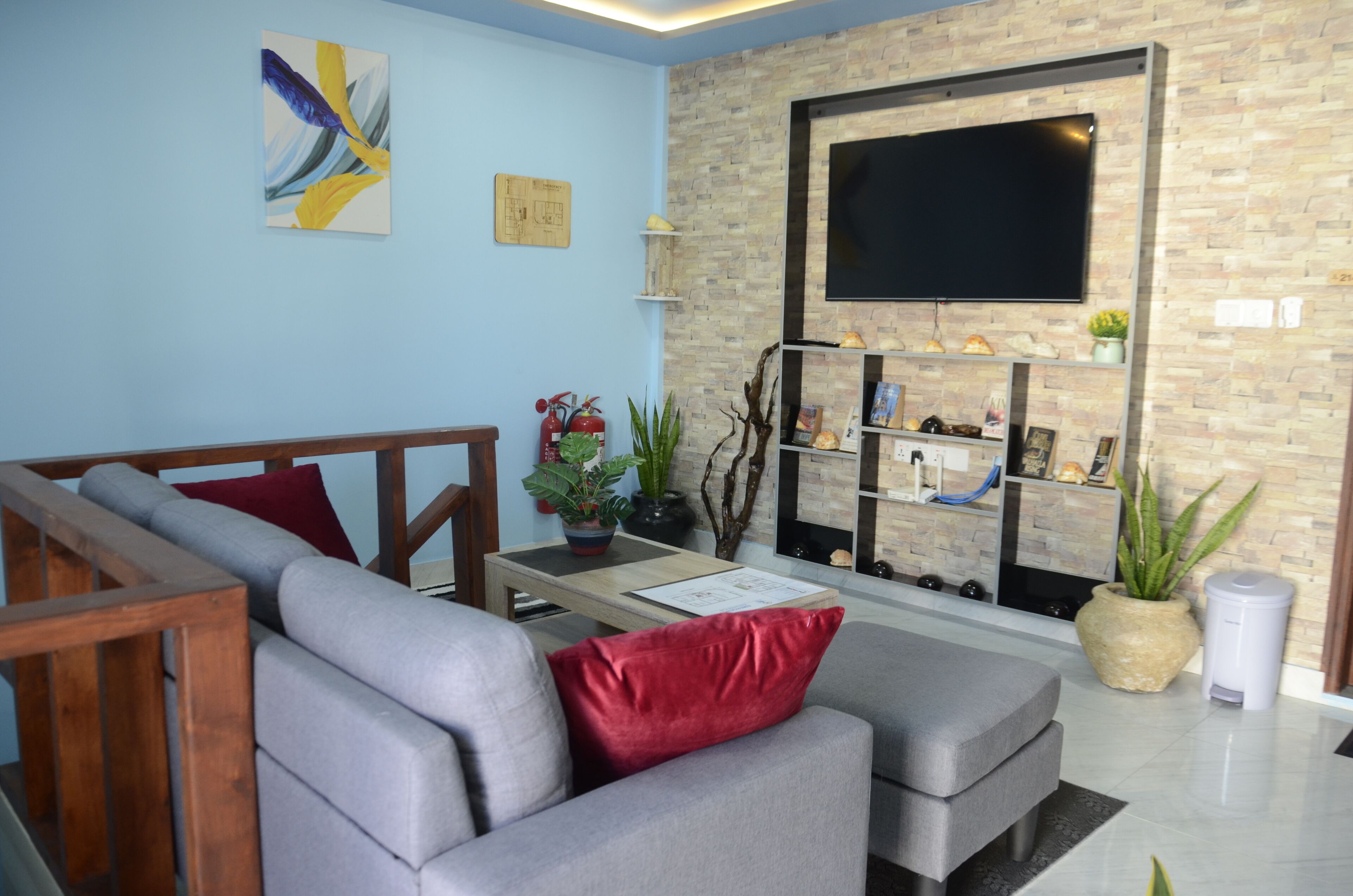 A living room with a grey sofa, red cushions, a wooden coffee table, a flat-screen TV mounted on a brick wall, and a small shelf with decorative items.