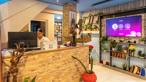 Un espace d’accueil avec un comptoir de réception en bois, un téléviseur affichant l’interface de YouTube, et des étagères ornées de plantes et d’objets décoratifs.