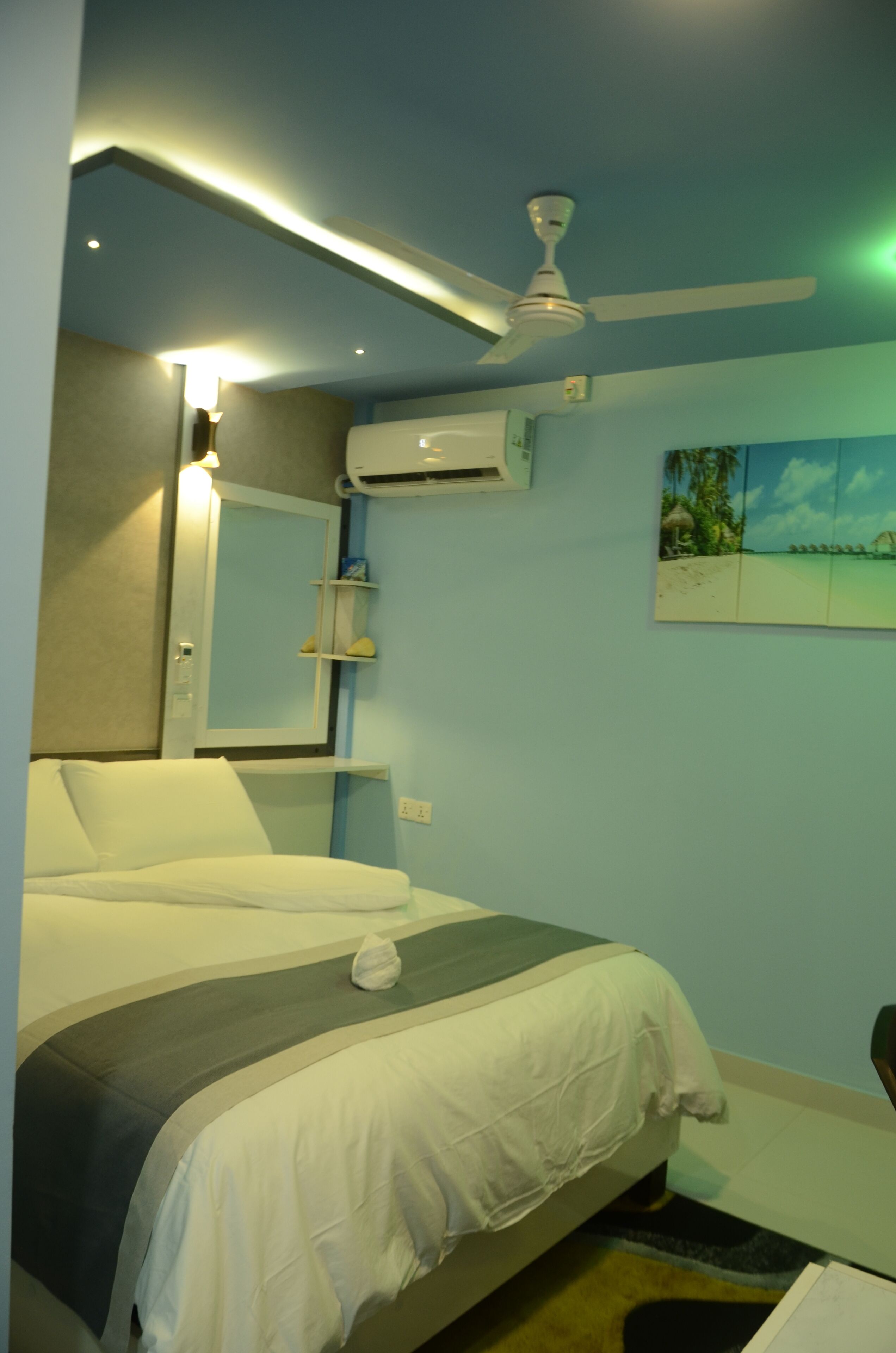 A hotel room with a bed, a ceiling fan, an air conditioning unit, a wall-mounted shelf with toiletries, and a beach-themed artwork.