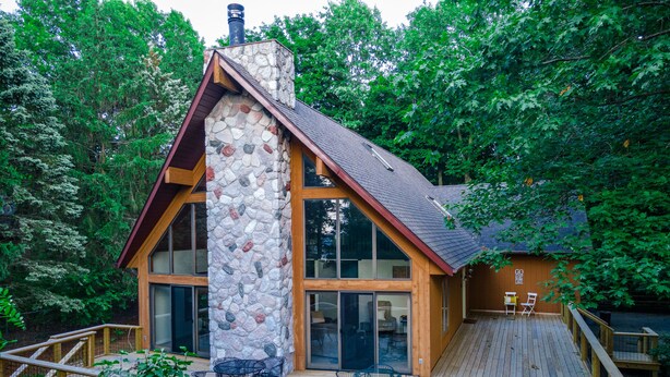 A cabin with a stone chimney, large windows, and a wooden deck with outdoor seating.