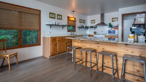 A modern kitchen with wooden cabinets, a large island with stools, and a view of greenery outside.