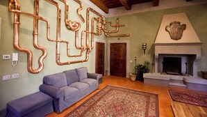 A living room with a grey sofa, patterned rugs, exposed copper pipes on the wall, a fireplace, and wooden beams on the ceiling.