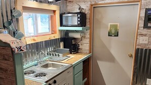 A compact kitchen with a sink, stove, and microwave. It features wooden walls and a door with a green sign. - Teeny Home in the Black Hills! (Custer)