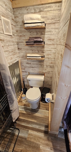 A compact bathroom with a rustic wooden wall, a white toilet, and multiple towel racks. - Not available (Custer)