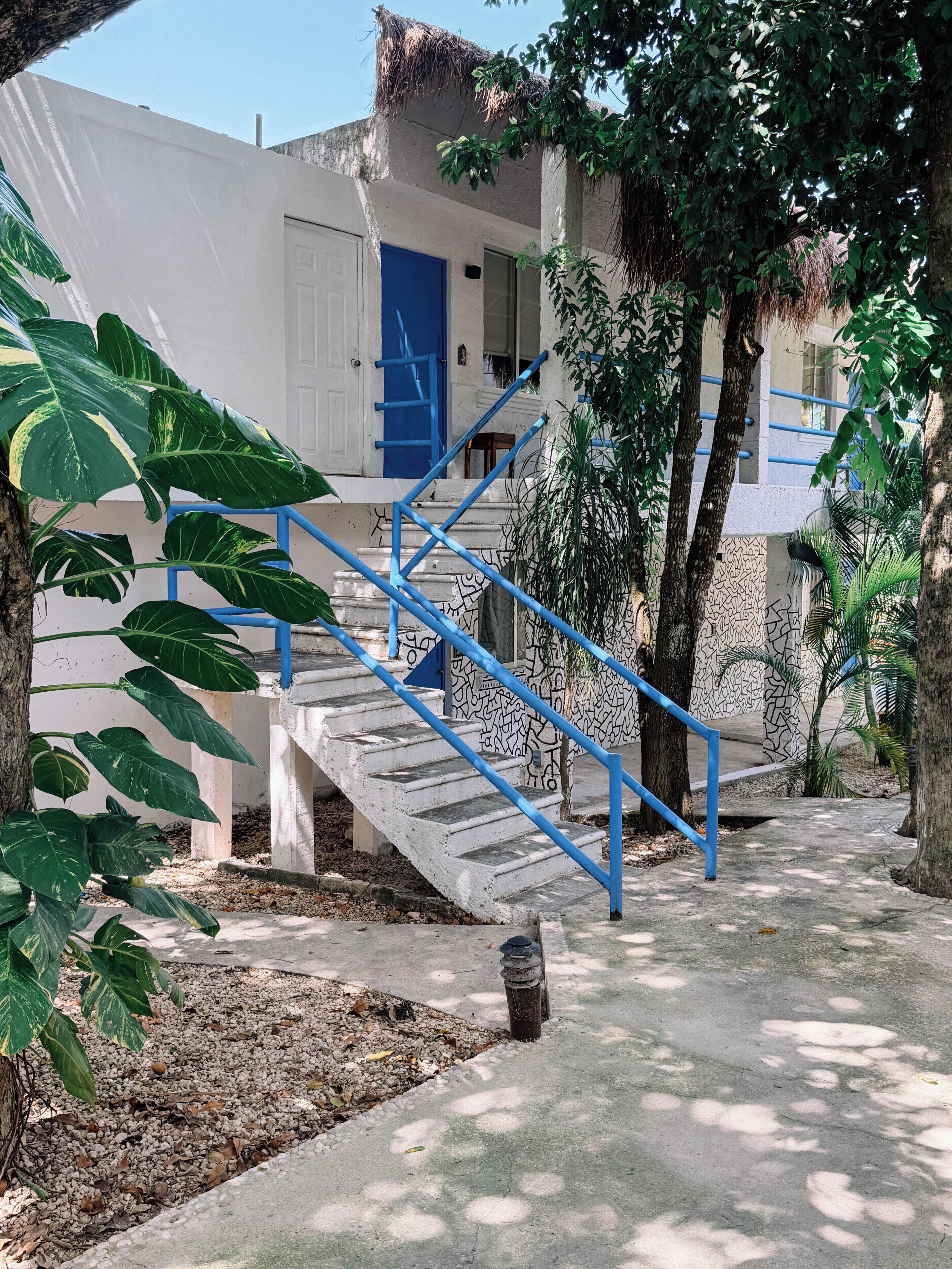 Un escalier en béton avec des rampes bleues mène à un bâtiment blanc doté d’une porte bleue.