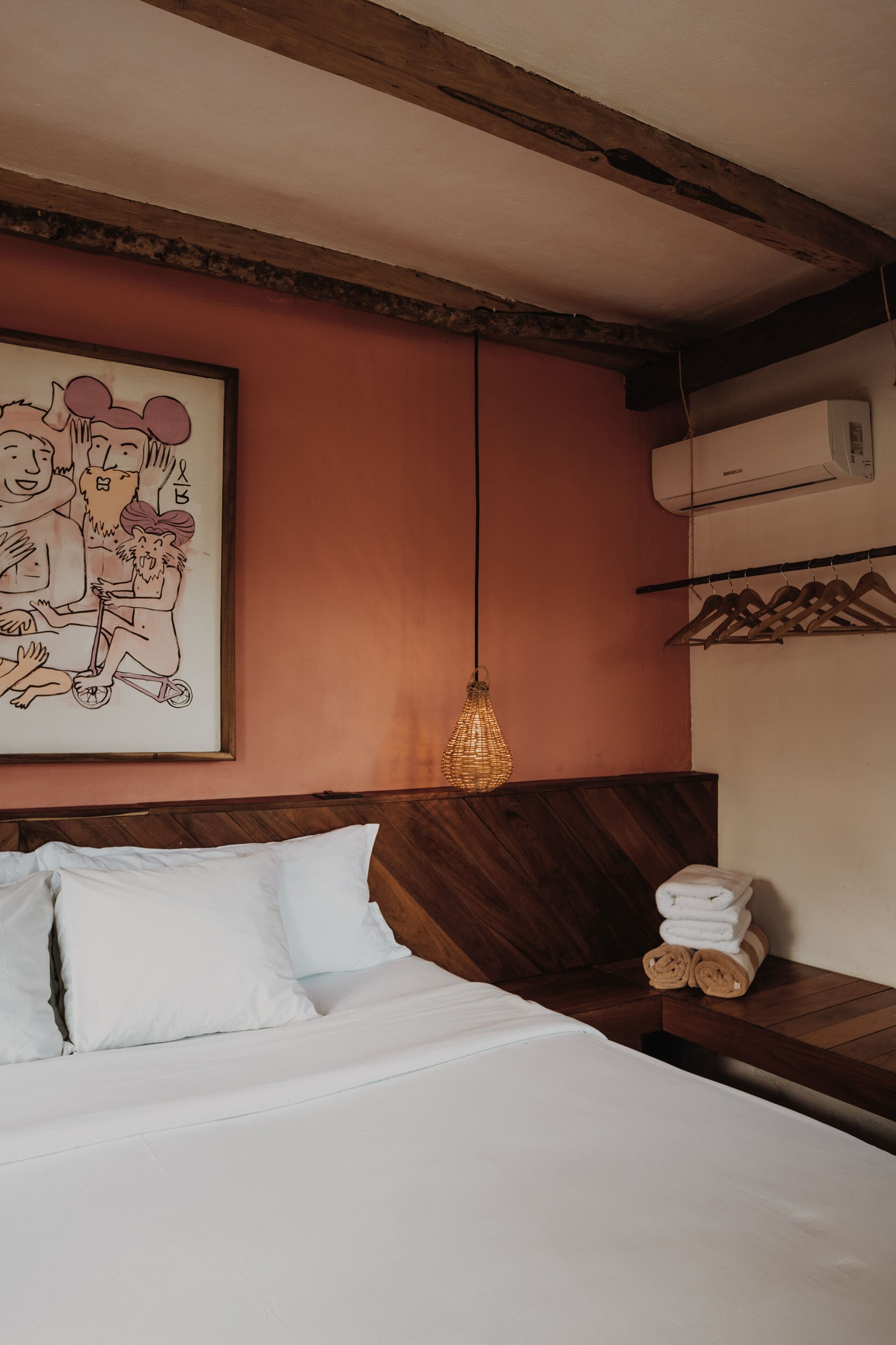Quarto de hotel com cama, cabeceira de madeira, obra de arte de desenho emoldurada, luminária suspensa e porta-toalhas com toalhas.