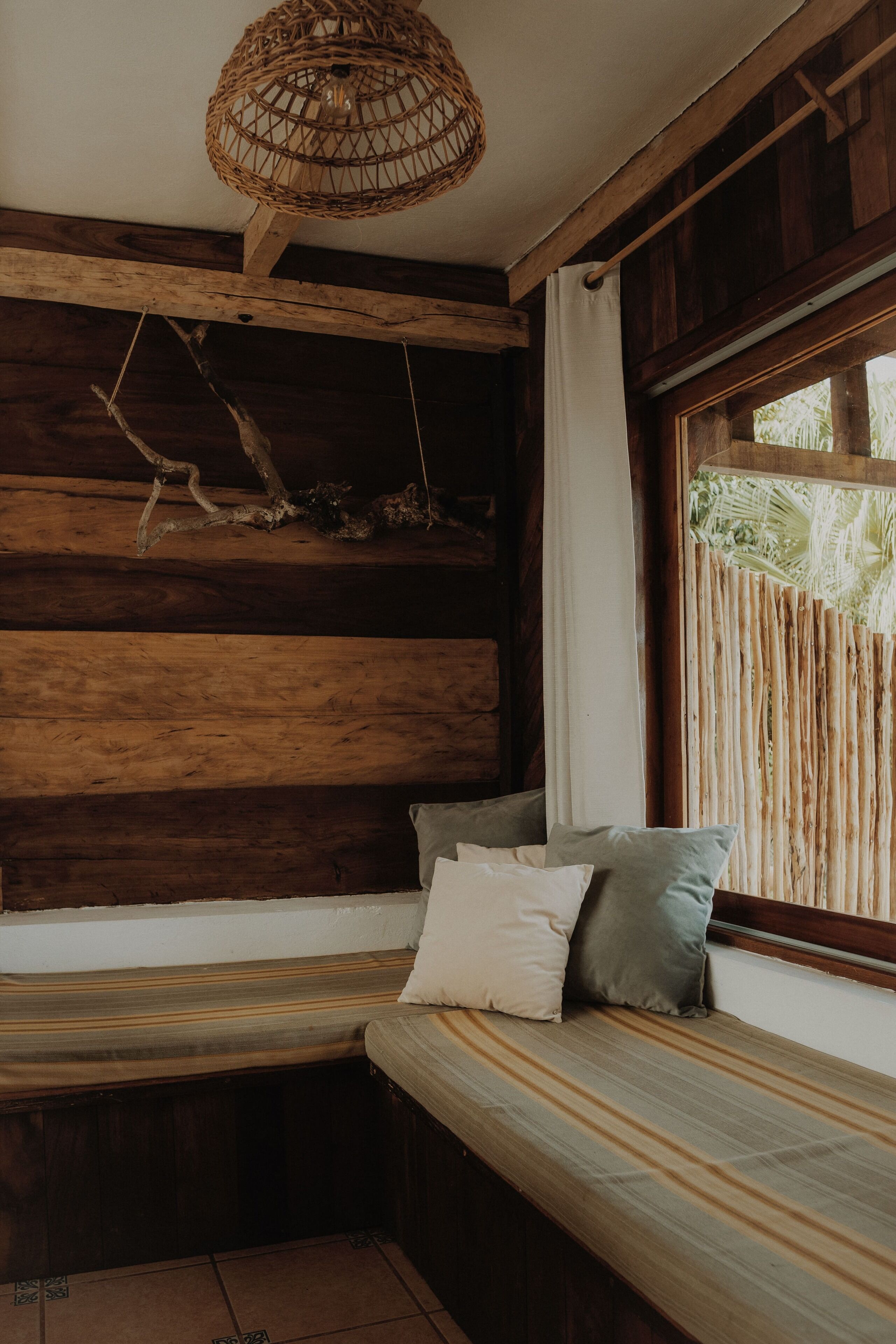 A wooden bench with cushions, a woven ceiling light, and a window with a curtain.