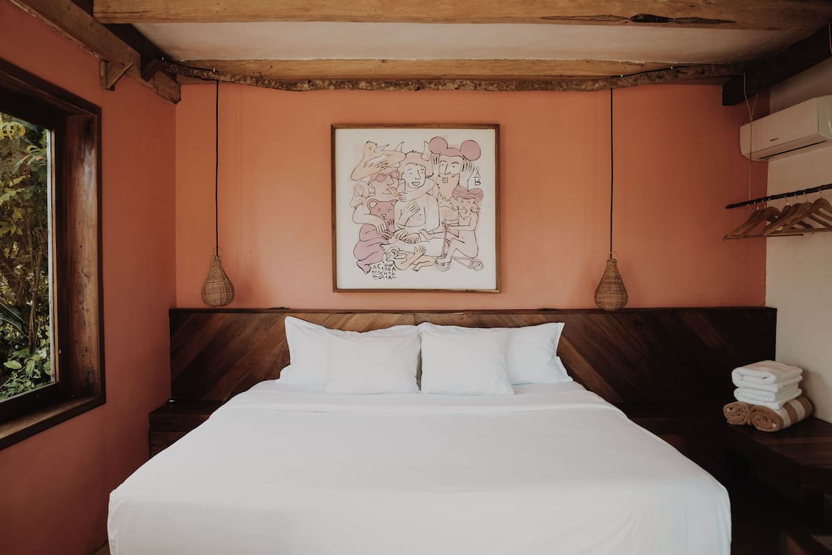 Una cama bien hecha con ropa de cama blanca, una ilustración caricaturesca enmarcada en la pared y un cabecero de madera.