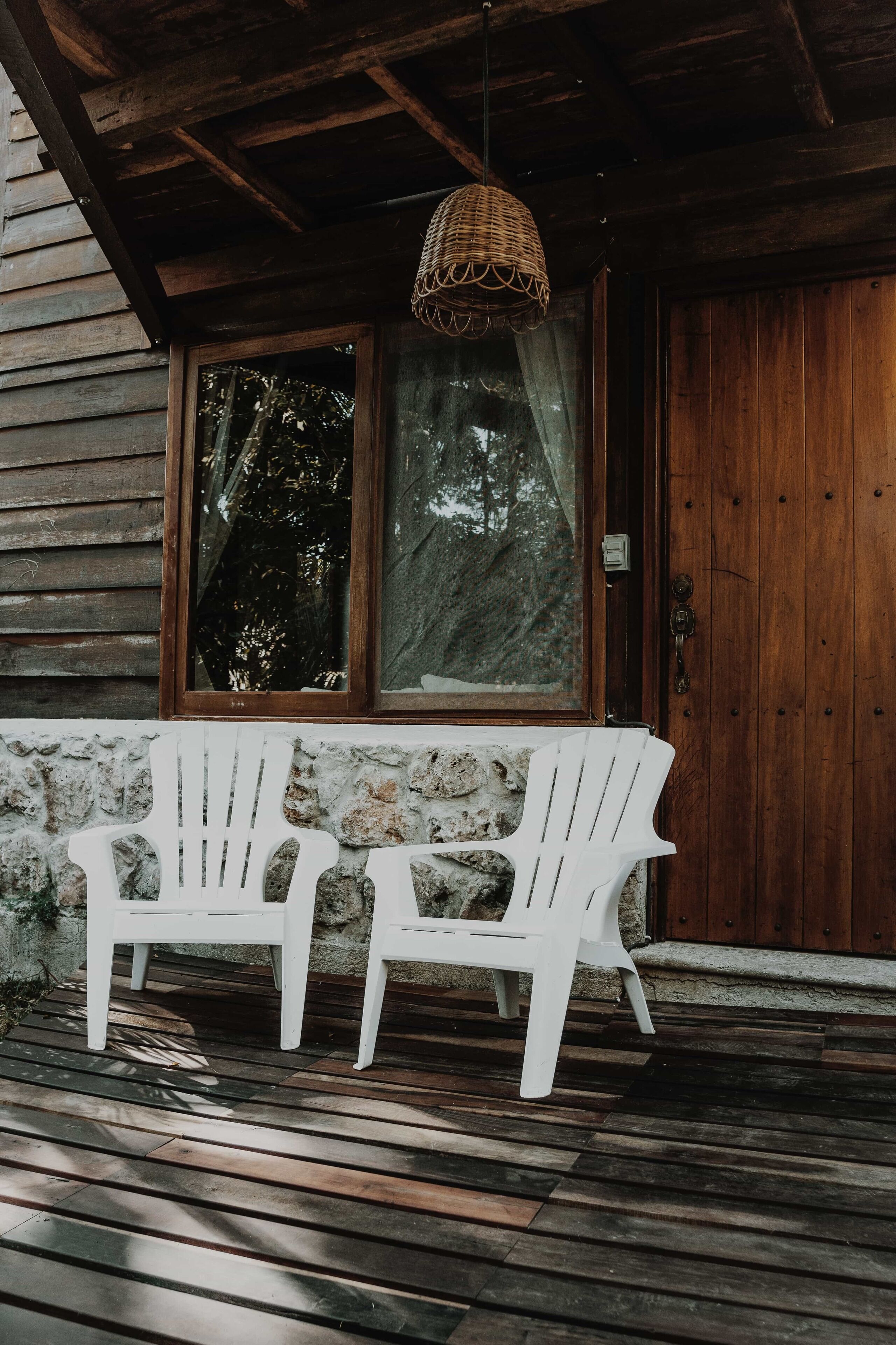 Twee witte Adirondack-stoelen op een houten terras voor een houten blokhut met een groot raam en een geweven lamp.