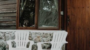 Twee witte Adirondack-stoelen op een houten terras voor een houten blokhut met een groot raam en een geweven lamp.
