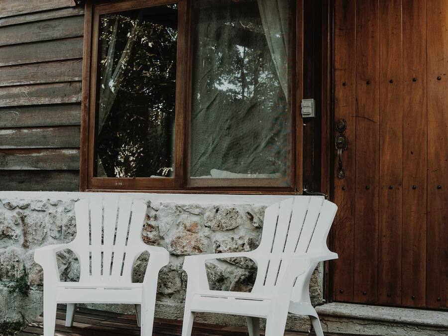 Twee witte Adirondack-stoelen op een houten terras voor een houten blokhut met een groot raam en een geweven lamp.