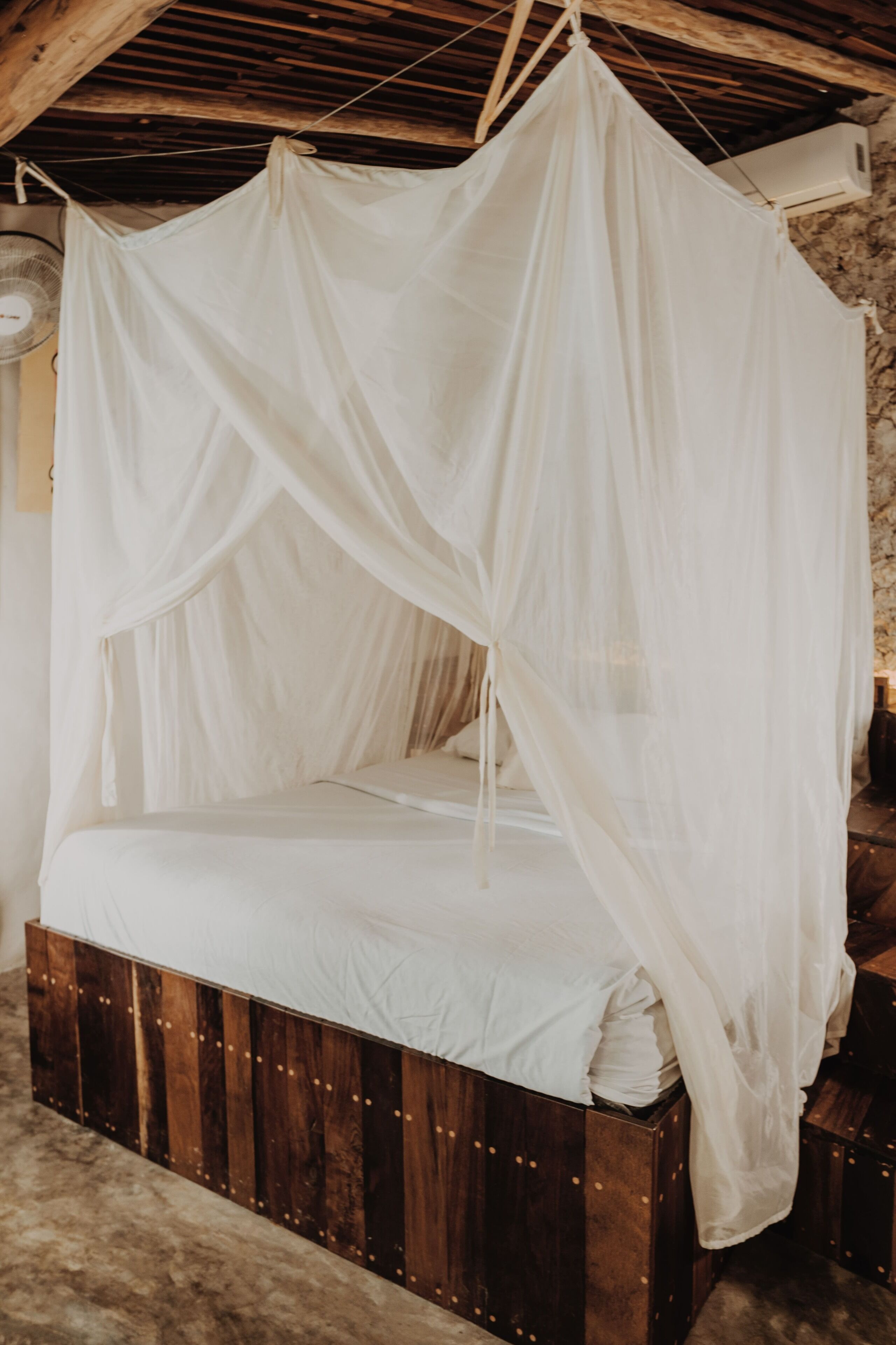 A four-poster canopy bed with a wooden base, set in a rustic room with exposed beams and a stone wall.