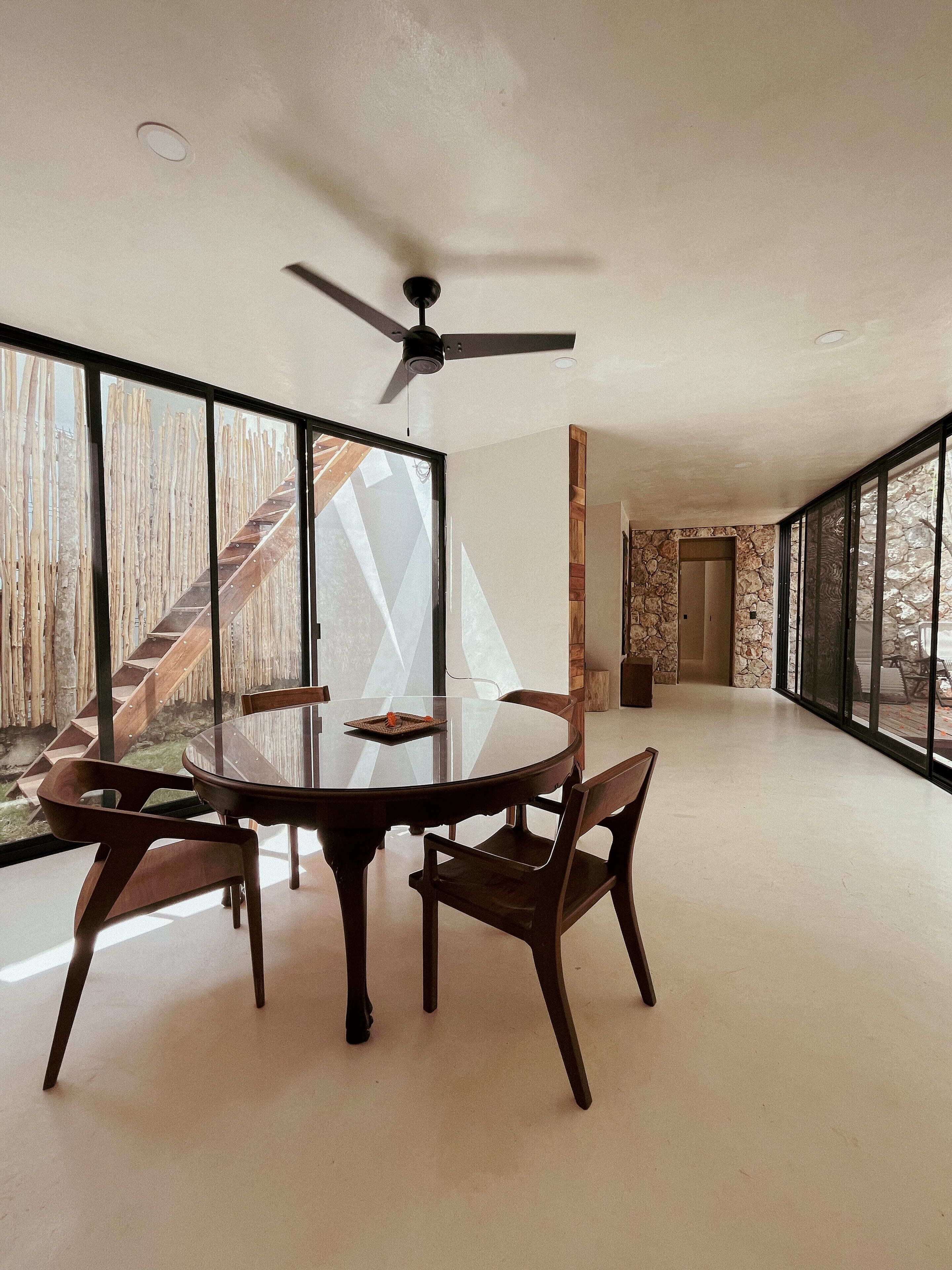 Una habitación amplia con una mesa redonda de vidrio, cuatro sillas de madera, un ventilador de techo y ventanas grandes que ofrecen vistas de una escalera de madera y un espacio exterior.