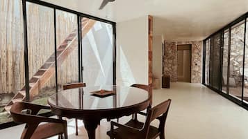 Una habitación amplia con una mesa redonda de vidrio, cuatro sillas de madera, un ventilador de techo y ventanas grandes que ofrecen vistas de una escalera de madera y un espacio exterior.