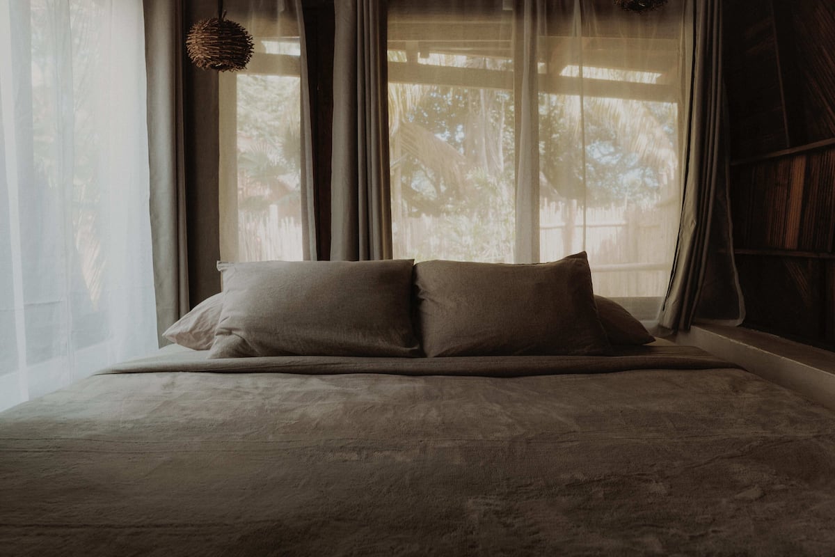 Una cama con colcha en tonos neutros y dos almohadas, frente a una ventana con cortinas translúcidas y una lámpara colgante de fibras naturales.