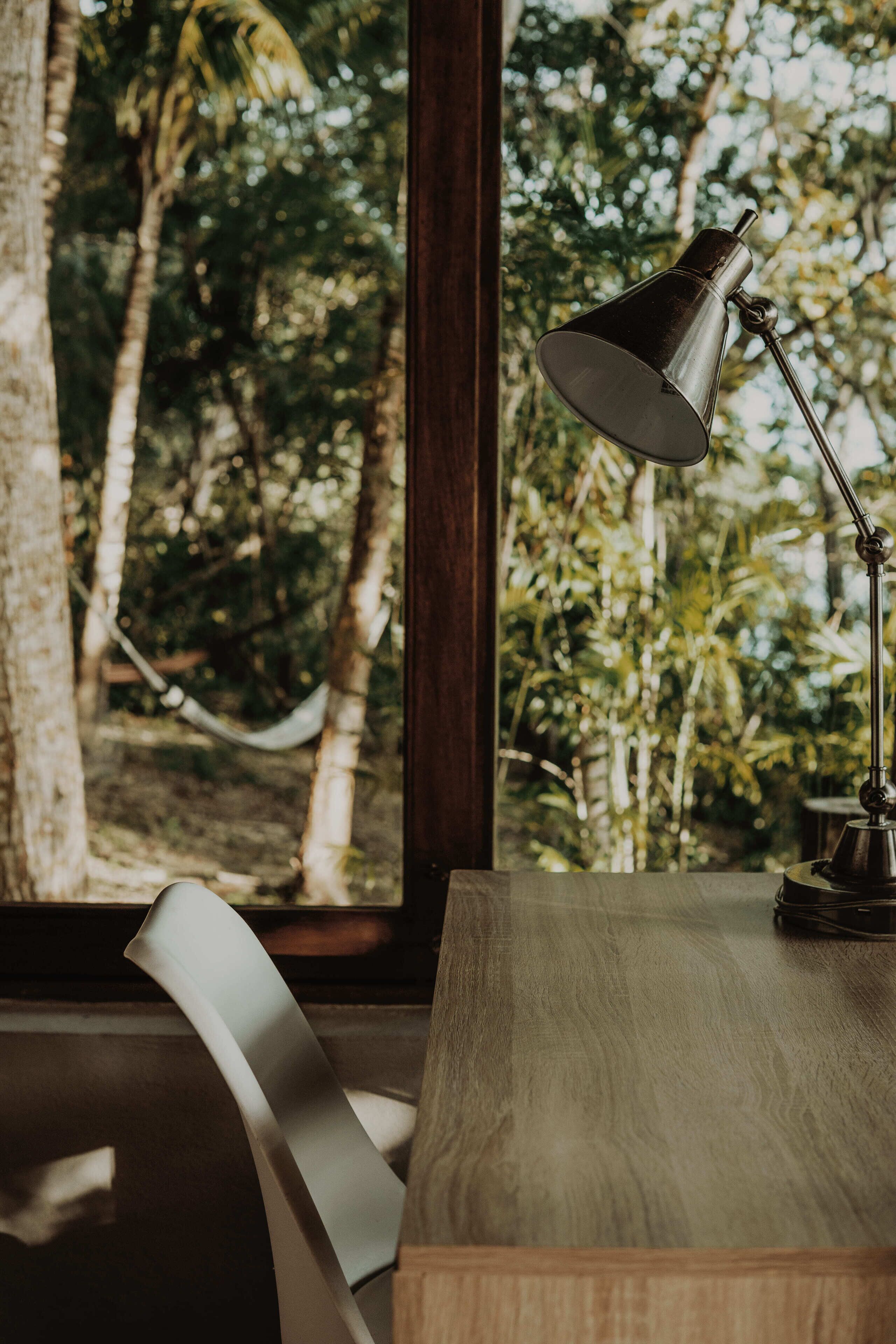 A desk with a lamp, a chair, and a view of trees through a window.