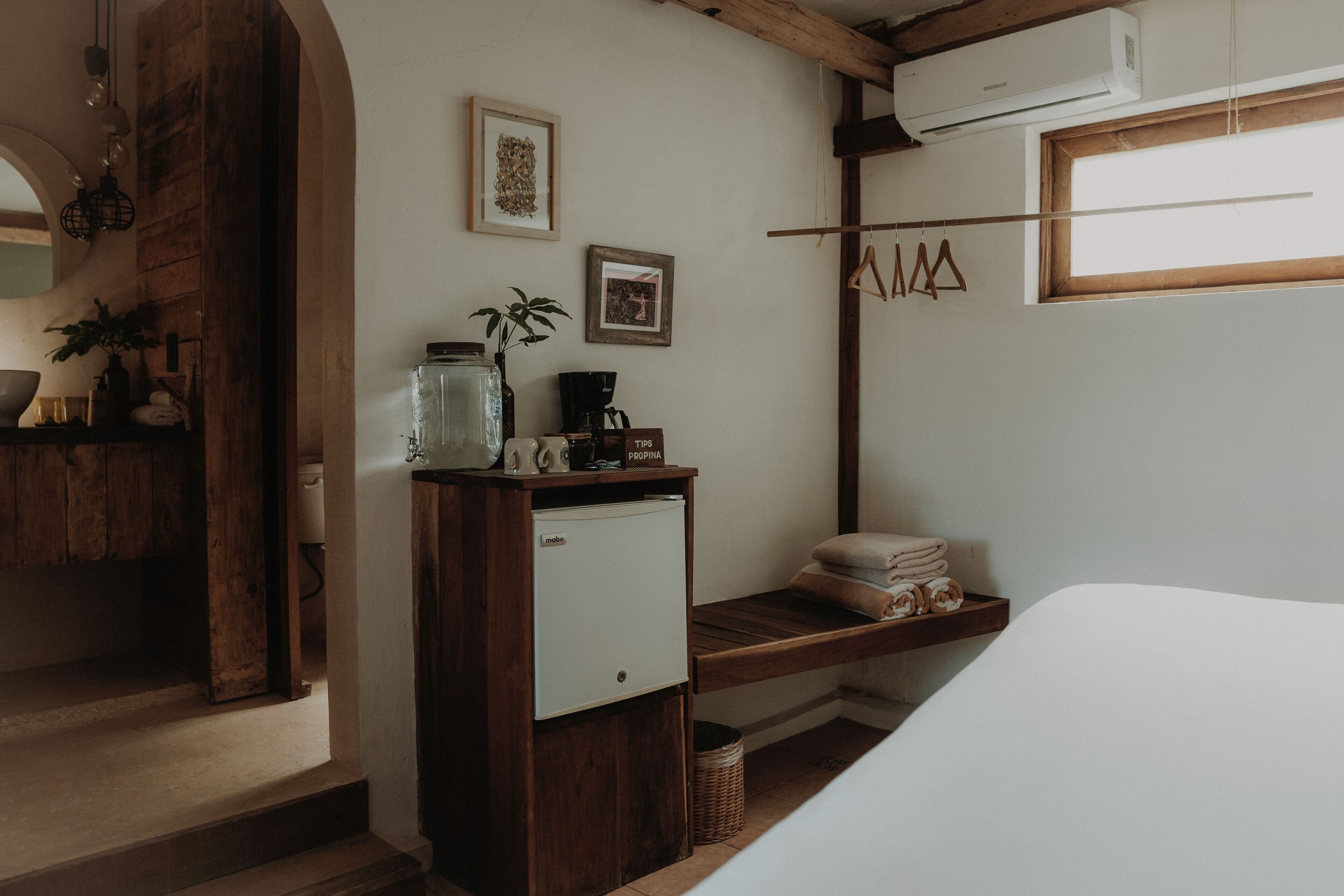 A bedroom with a bed, a small white refrigerator, a shelf with towels, a hanging clothes rack, and a window with a wooden frame.