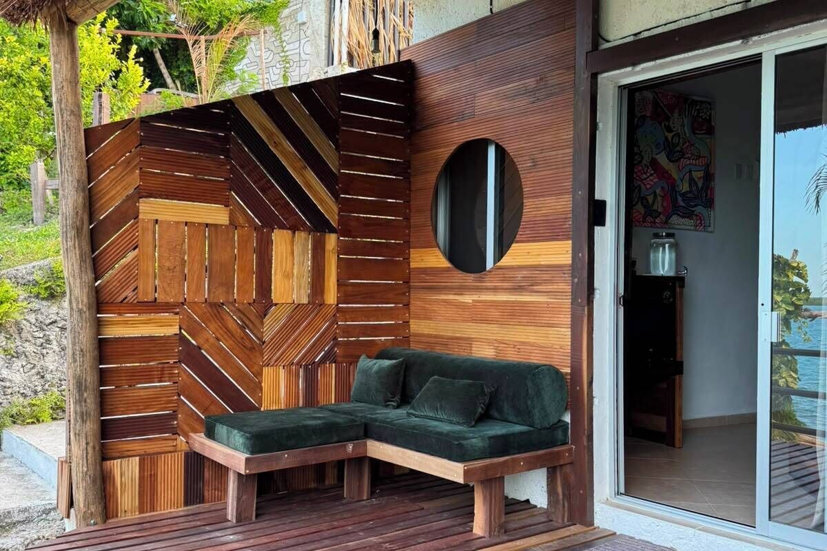 A modern outdoor seating area with a wooden bench and a sofa, a circular mirror, and a view of the sea.