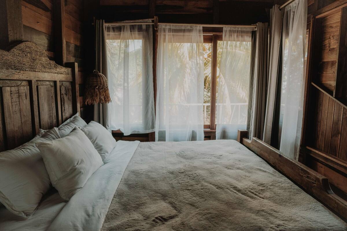 Una habitación estilo cabaña de madera con una cama grande, cortinas translúcidas y una cesta tejida colgando junto a la ventana.