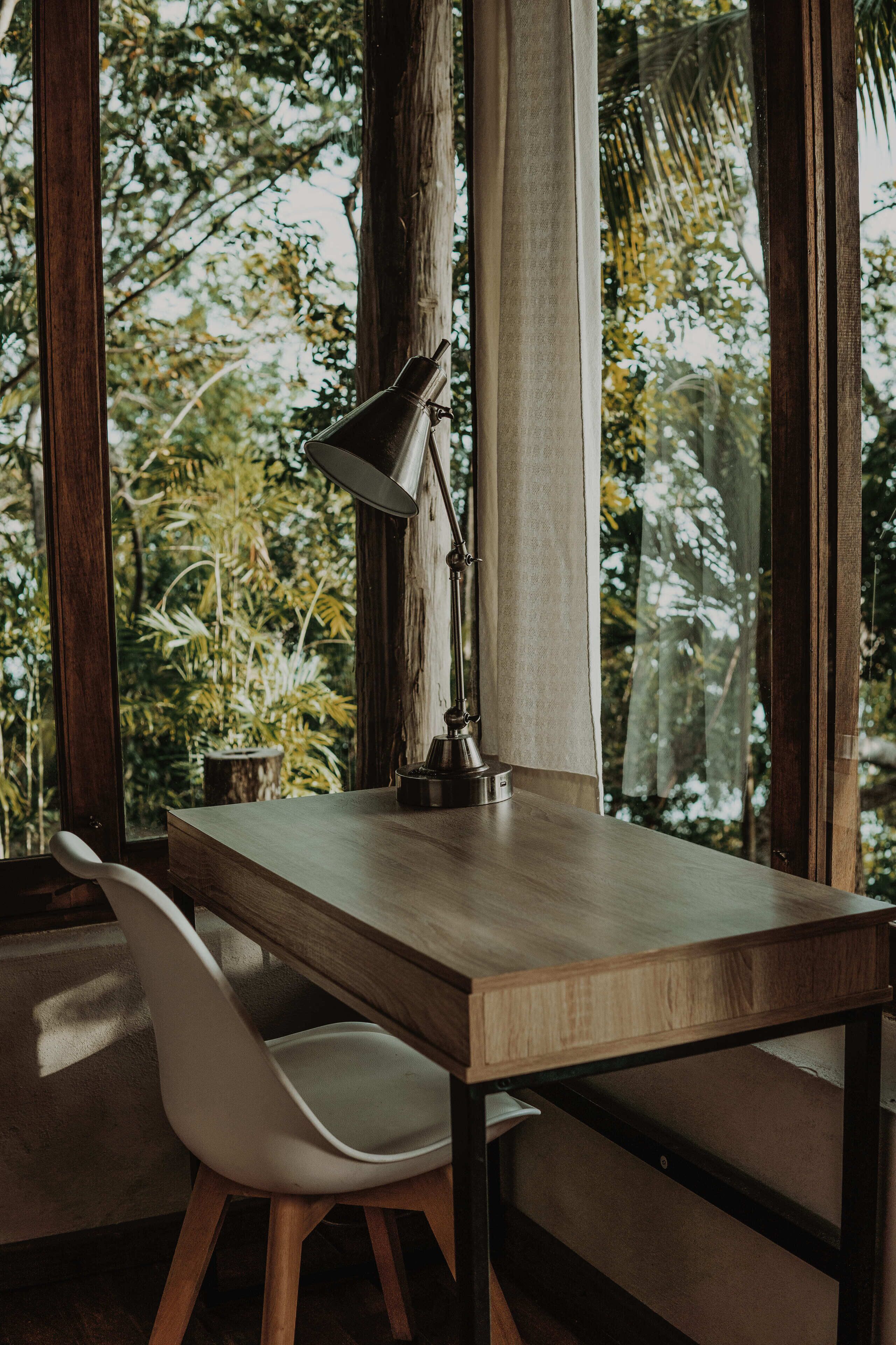 A wooden desk with a lamp, a white chair, and a view of trees through large windows.