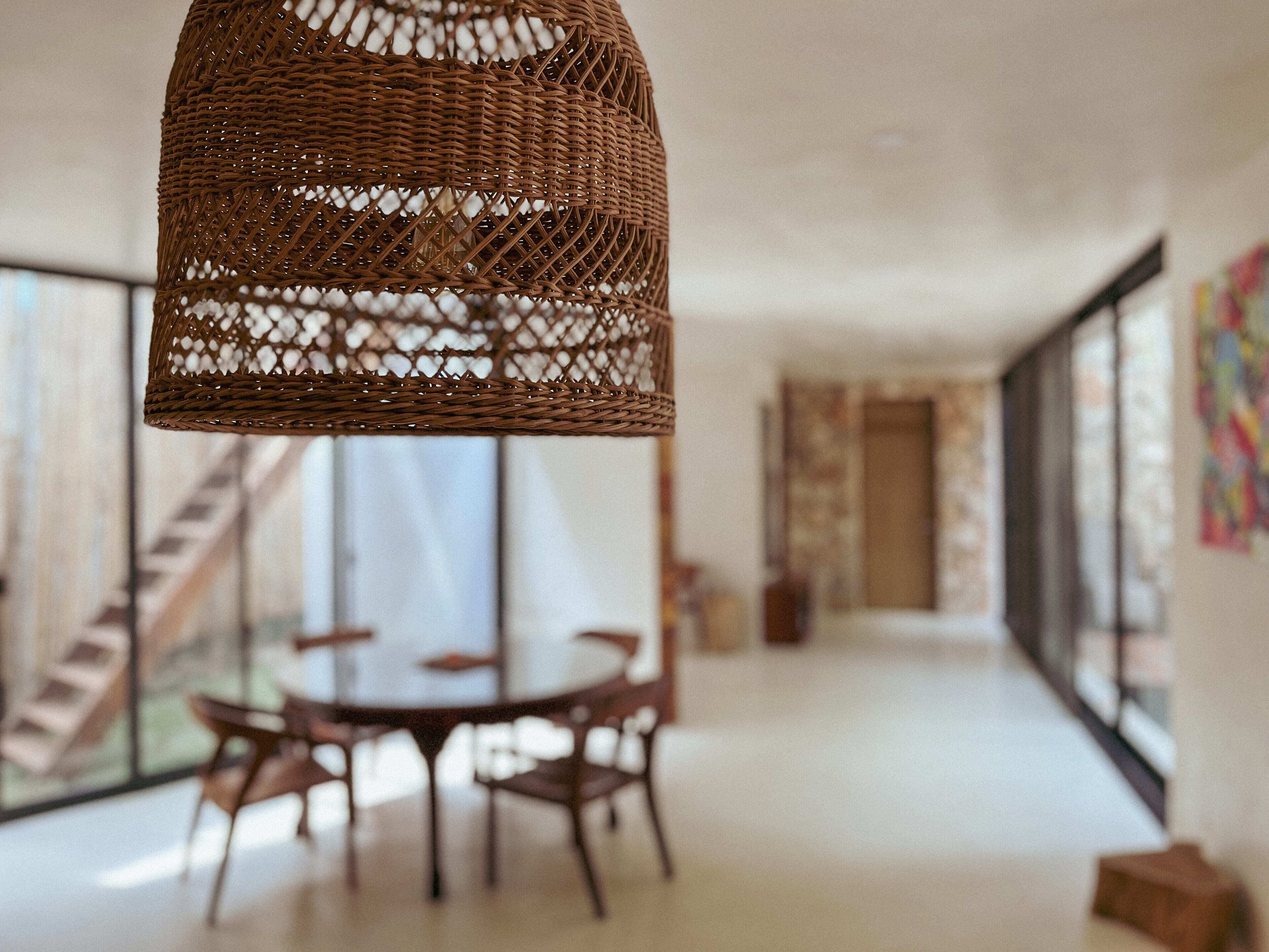 Un intérieur moderne agrémenté d’un luminaire suspendu en osier, d’une table de salle à manger ronde avec des chaises, et de grandes fenêtres offrant une vue sur l’extérieur.