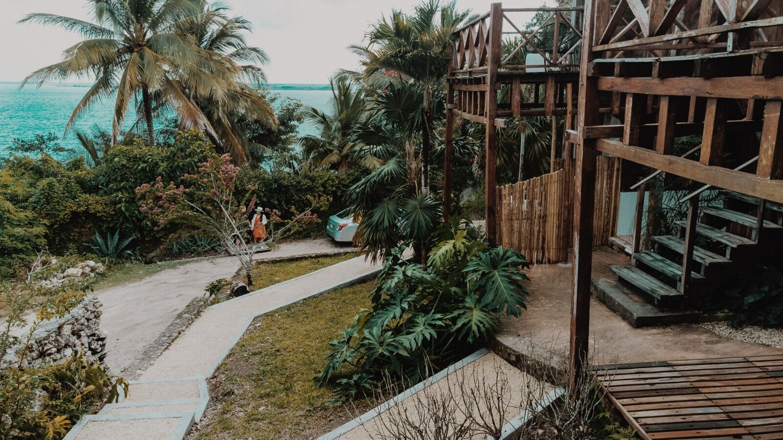 Una terraza de madera con una escalera que lleva a una construcción de madera, rodeada de vegetación tropical y vista al océano.