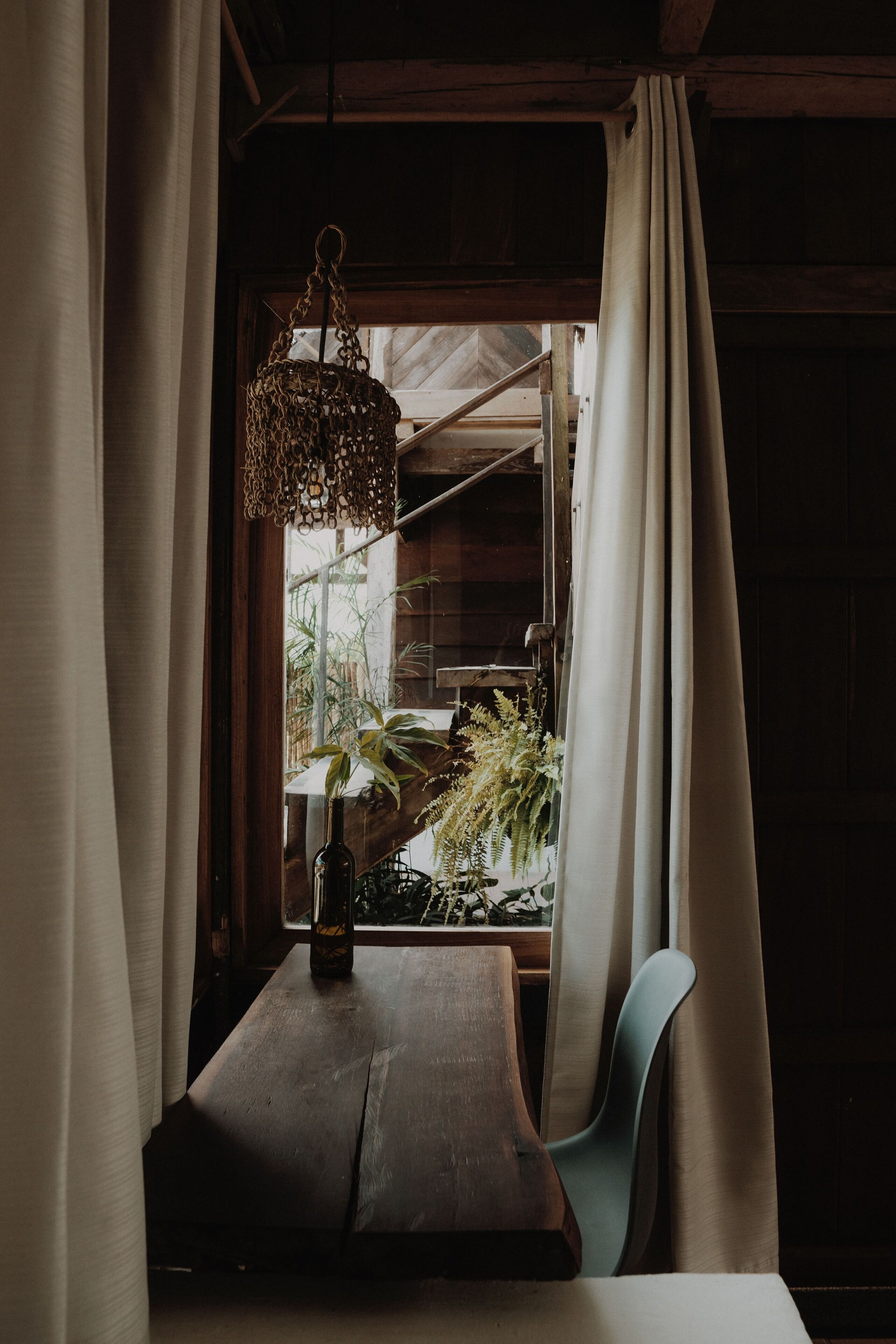 A wooden table with a bottle and a plant, a chair, and a hanging light fixture.
