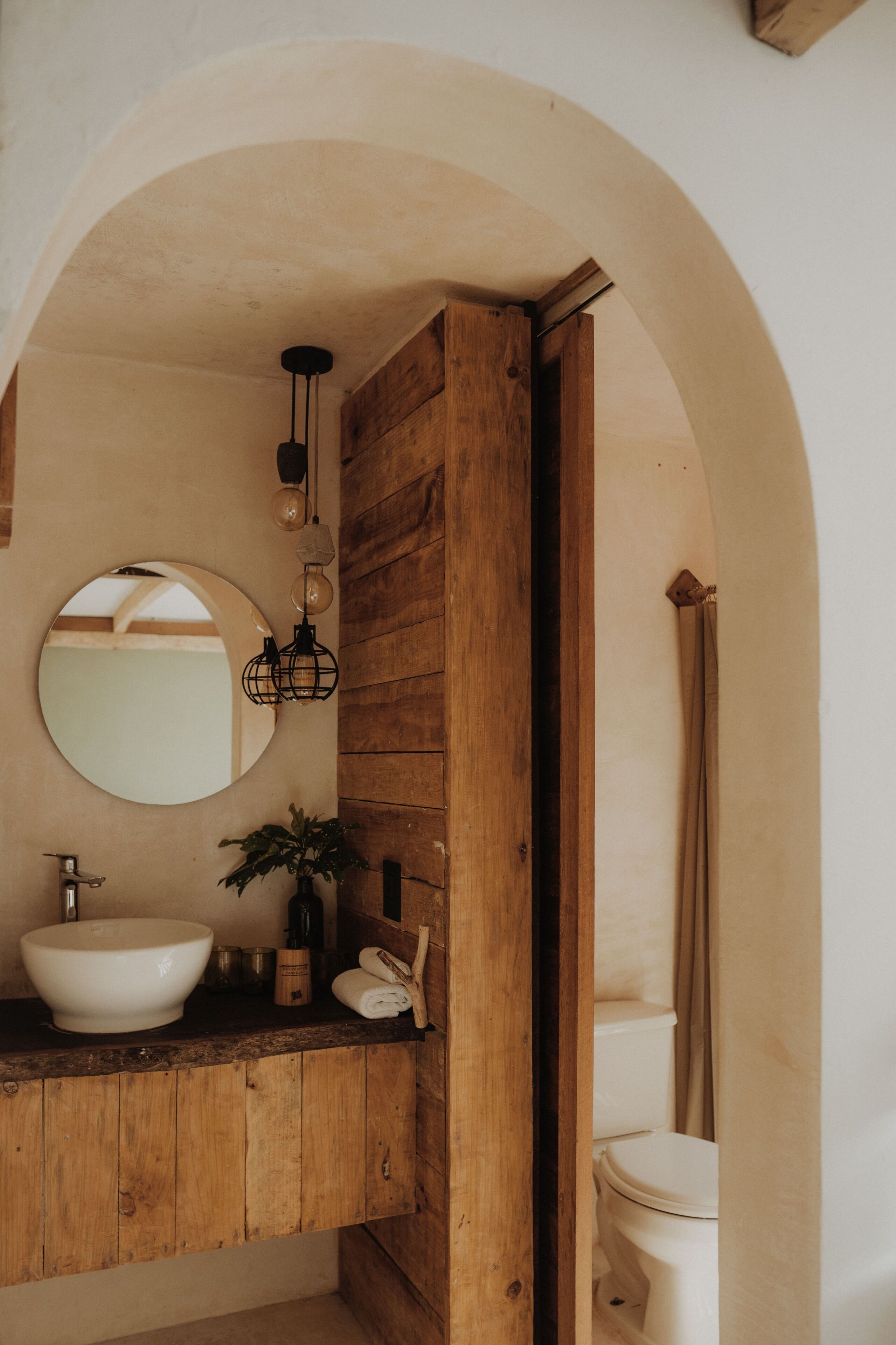 Une salle de bain avec un meuble-lavabo en bois, un miroir rond, un lavabo et des toilettes.