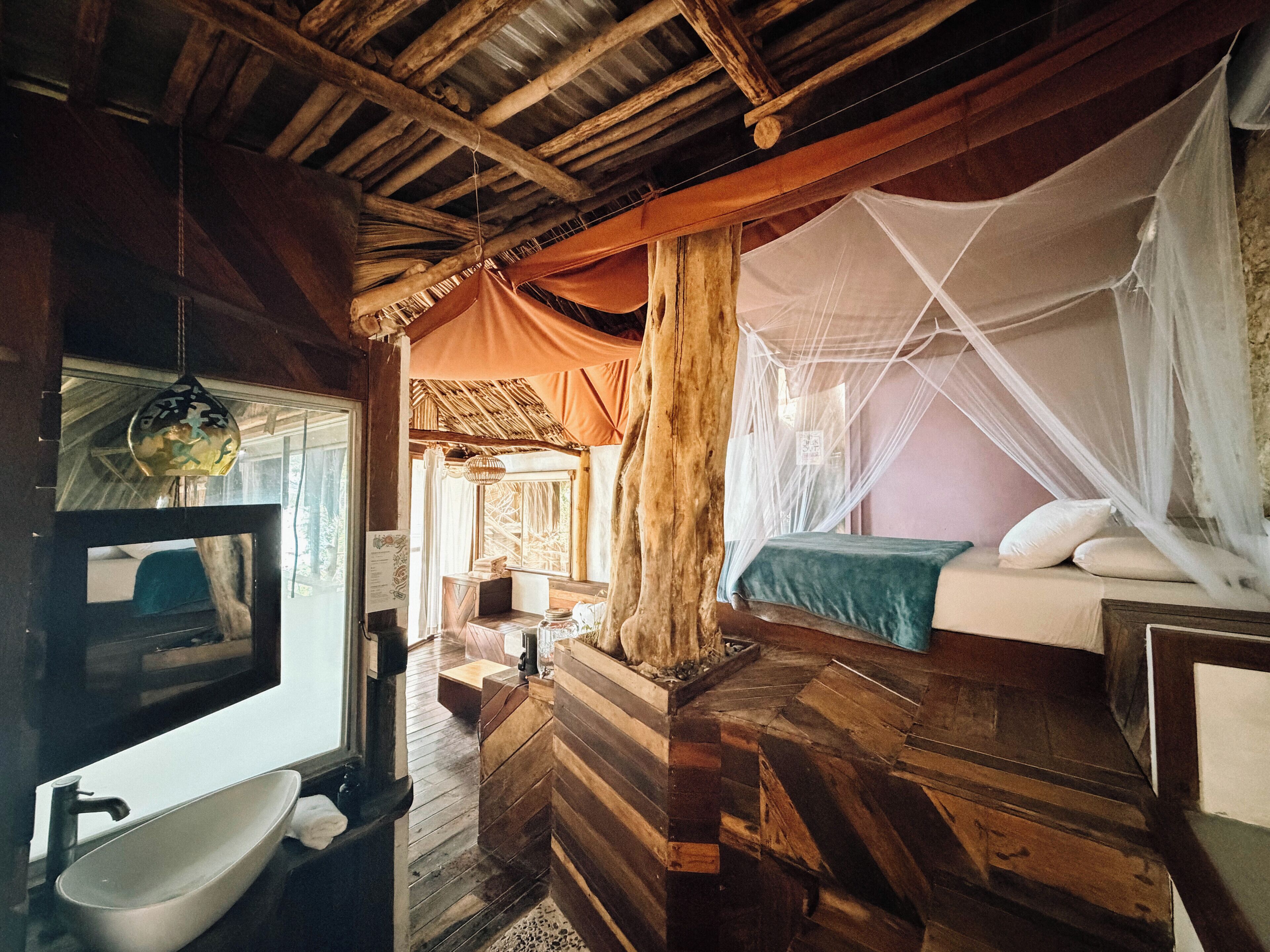 A rustic room with a bed, a wooden bench, a sink, and a window with curtains.