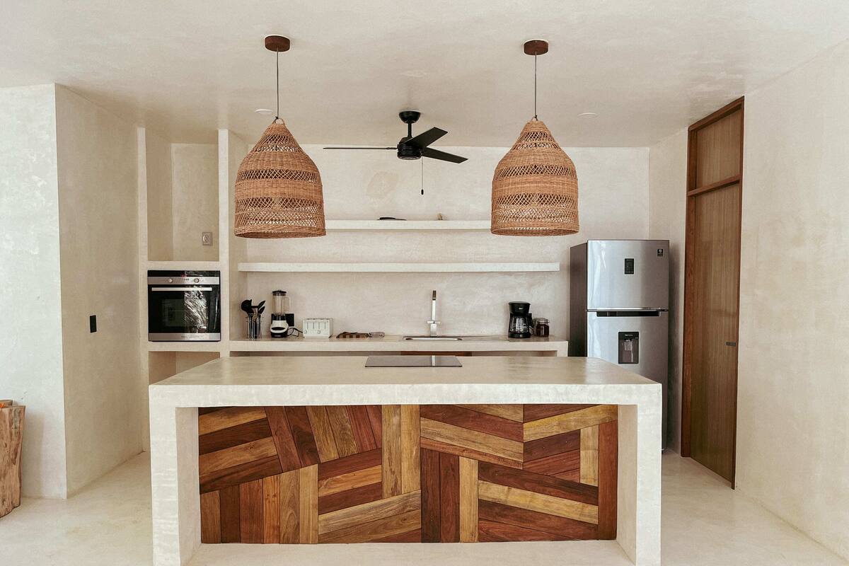 Una cocina moderna con una isla central, dos lámparas colgantes de mimbre, horno empotrado, refrigerador y ventilador de techo.