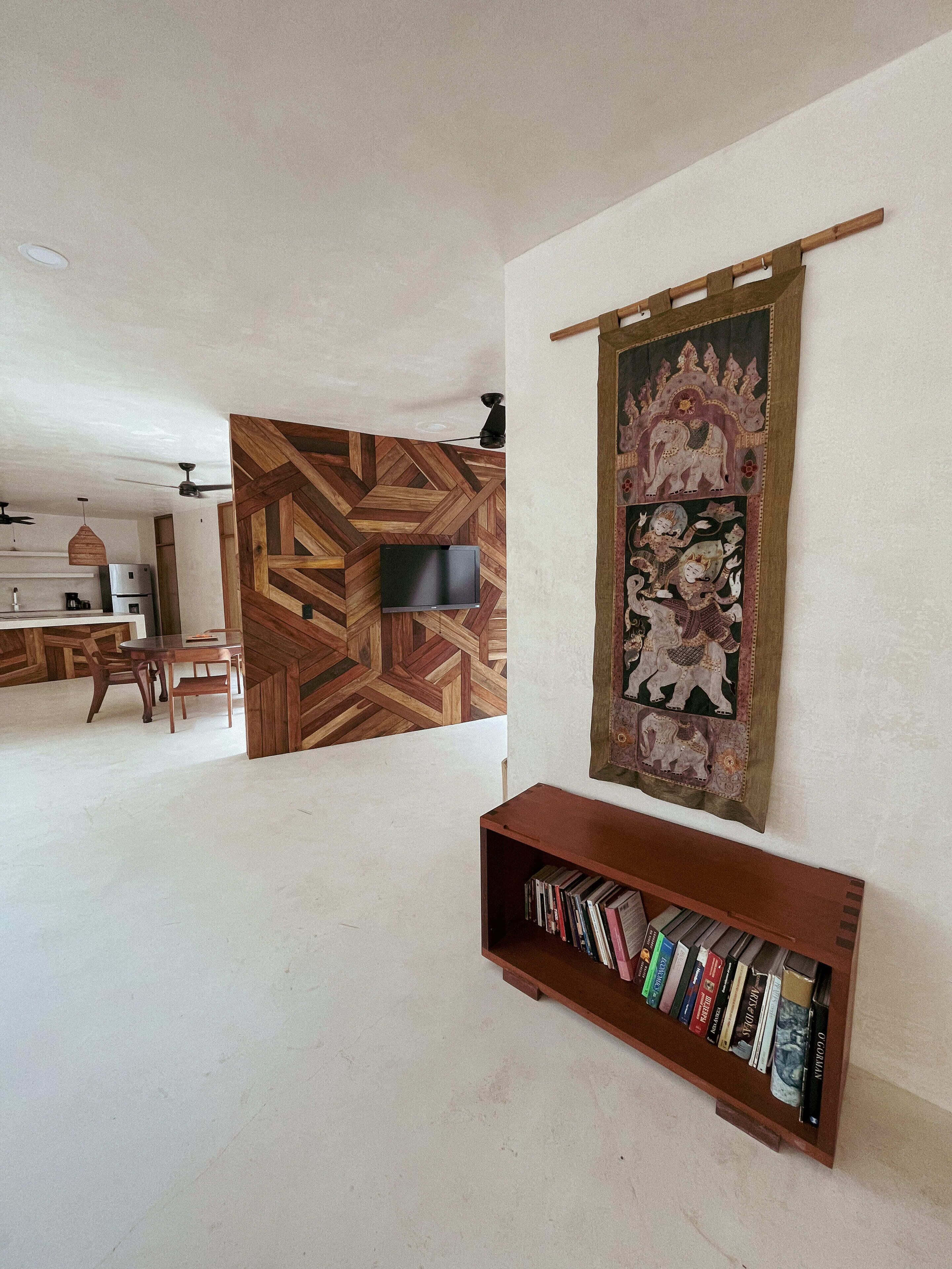 A living room with a wooden herringbone floor, a flat-screen TV mounted on a wooden panel, a bookshelf filled with books, and a decorative tapestry with elephant motifs.