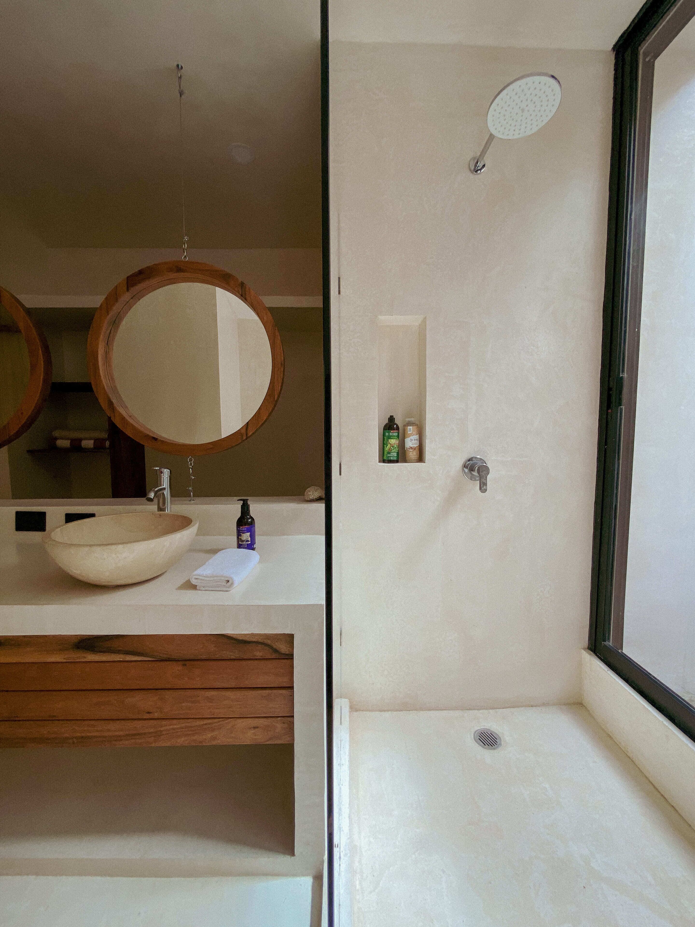 A modern bathroom with a wooden vanity, a large mirror, a round mirror, a beige sink, and a shower with a handheld showerhead.