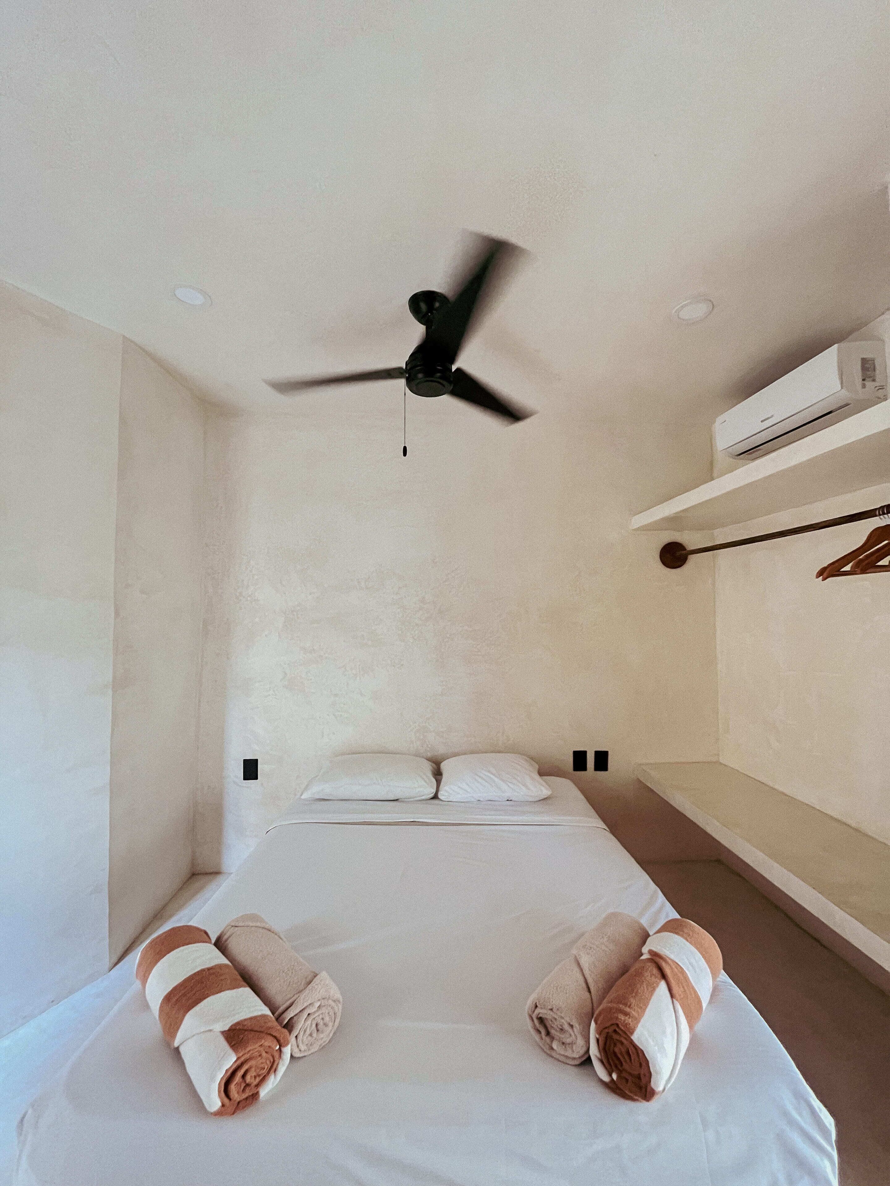 A hotel room with a bed, two rolled towels, a ceiling fan, and an air conditioning unit.