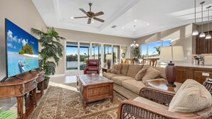 A spacious living room with a large flat-screen TV, comfortable seating, a ceiling fan, and a view of a pool through large glass doors. - Dockside Shore- Waterfront Home in Bayshore Arts District of Naples (Naples)