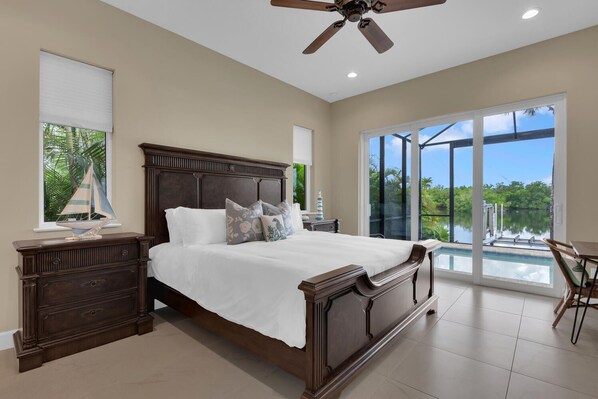 A spacious bedroom with a large bed, a ceiling fan, and a view of a pool and greenery.