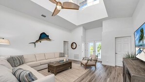 A spacious living room with a high ceiling, a ceiling fan, a large window, a sofa, a coffee table, a chair, and a television.
