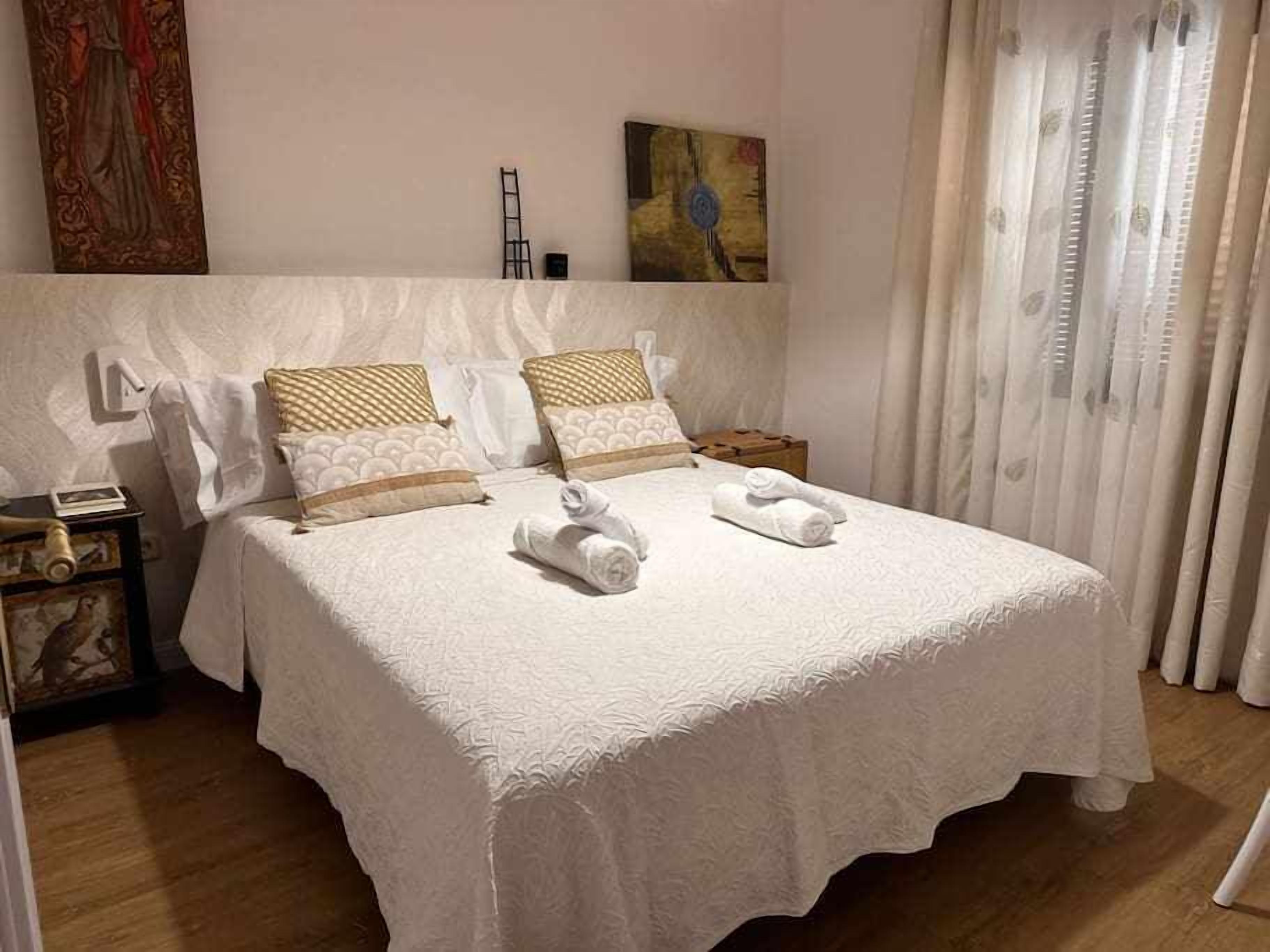 A neatly made bed with white linens and two rolled towels, a nightstand with a lamp, and a painting on the wall.