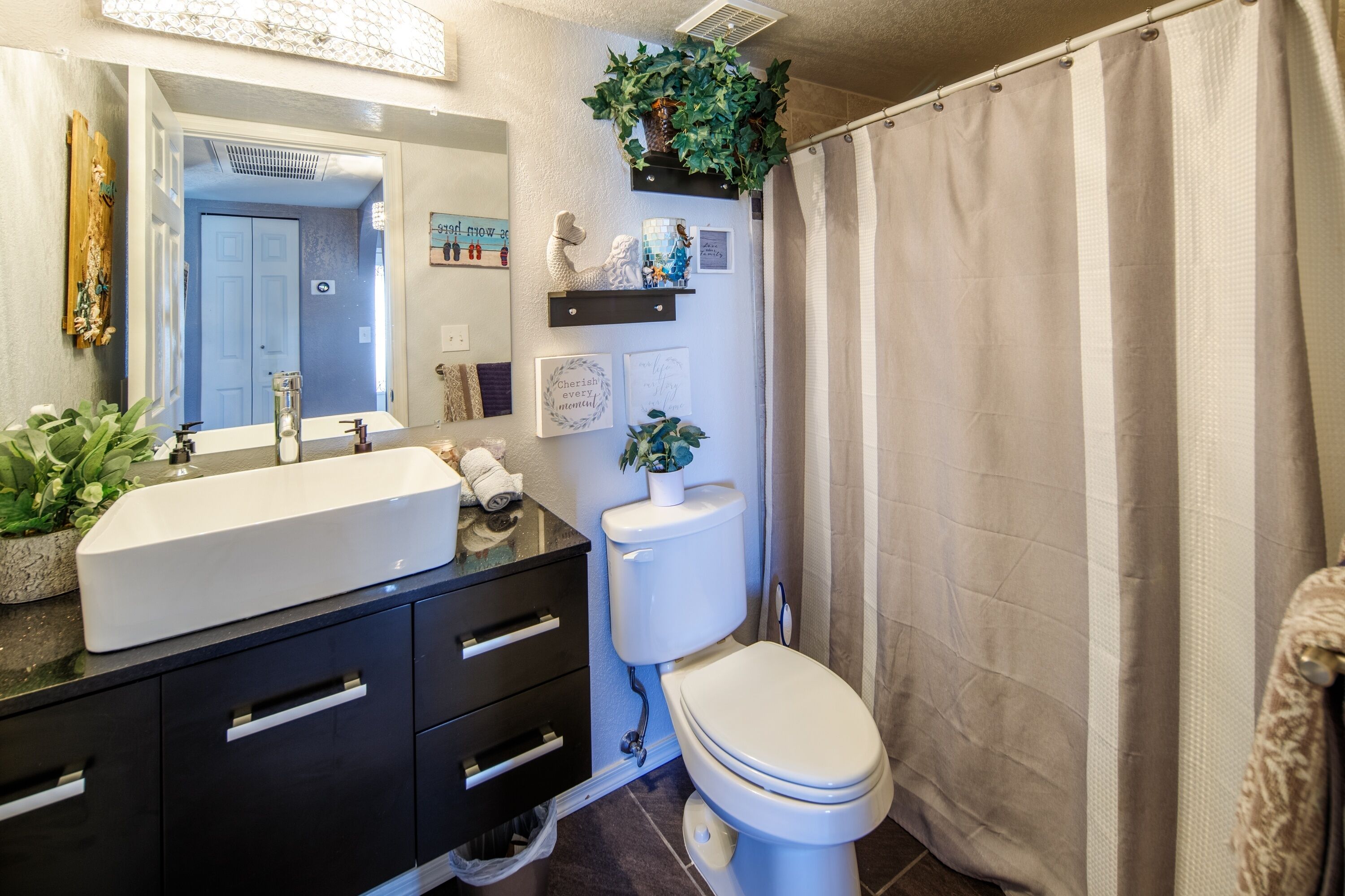 Et badeværelse med en hvid vask, et sort toiletbord og et toilet. Der er planter på toiletbordet, og der er et bruseforhæng.