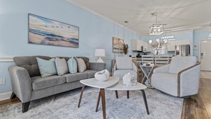 Un salon moderne avec un canapé gris, une table basse blanche et un fauteuil gris. Un tableau représentant des oiseaux est accroché au mur, et on aperçoit un coin cuisine au fond.
