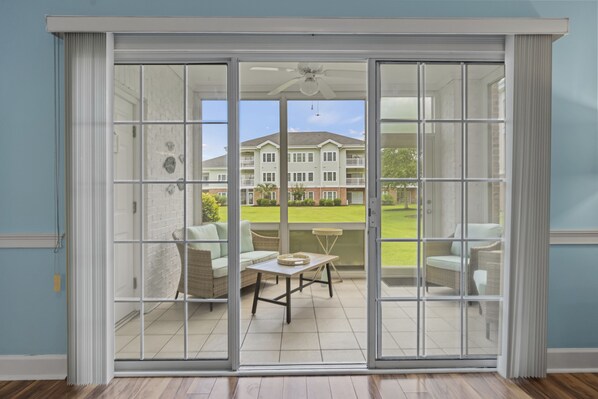 A room with a sliding glass door leading to an outdoor patio with a wicker sofa, a small table, and a view of a multi-story building in the background.