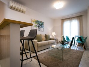 Een moderne woonkamer met een bank, twee barkrukken, een glazen salontafel en een eethoek met een tafel en stoelen.